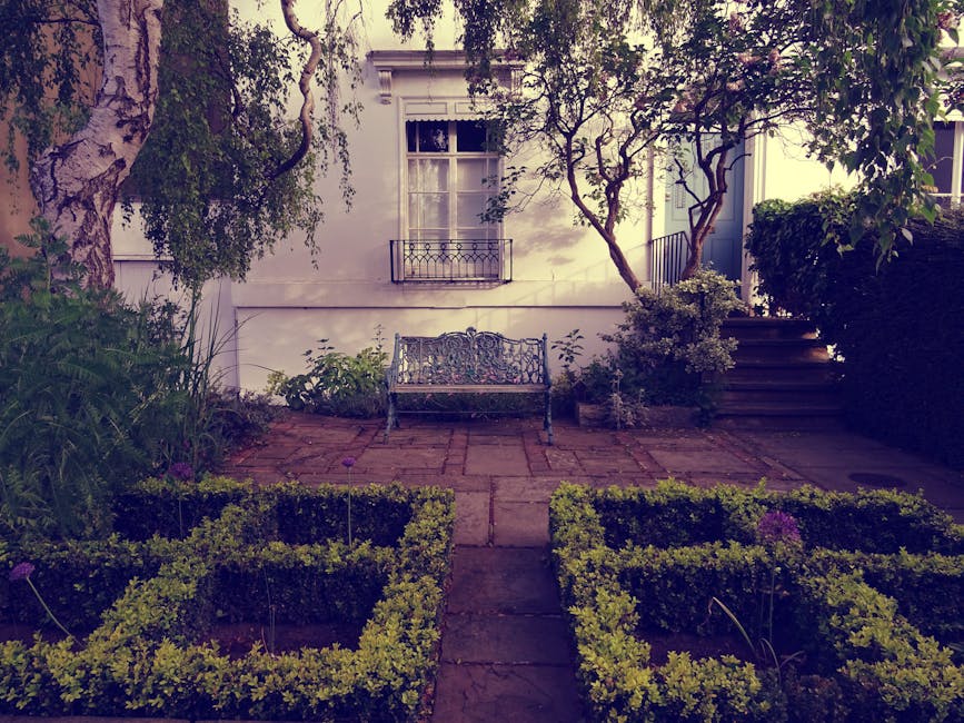 A small, well-maintained garden featuring a paved stone patio area with a decorative wrought iron bench positioned against a white exterior wall of a building, which has a window with white trim and a